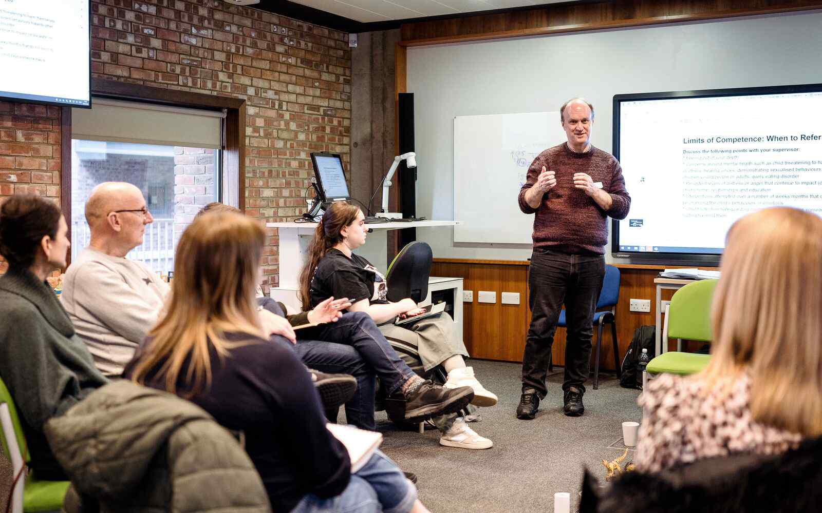 A classroom with a circle of people seated, facing the course leader who is stood teaching in front of a screen showing slides