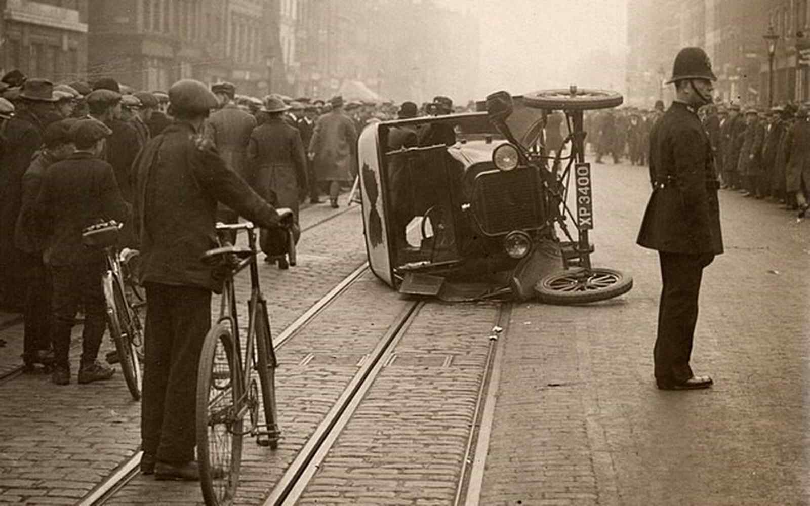 A police officer and crowd stand alongside a car which has been flipped onto it's side by protesters during the 1926 strike.