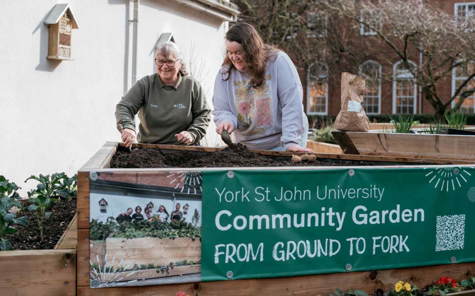 Two people gardening in raised wooden planters behind a community garden banner