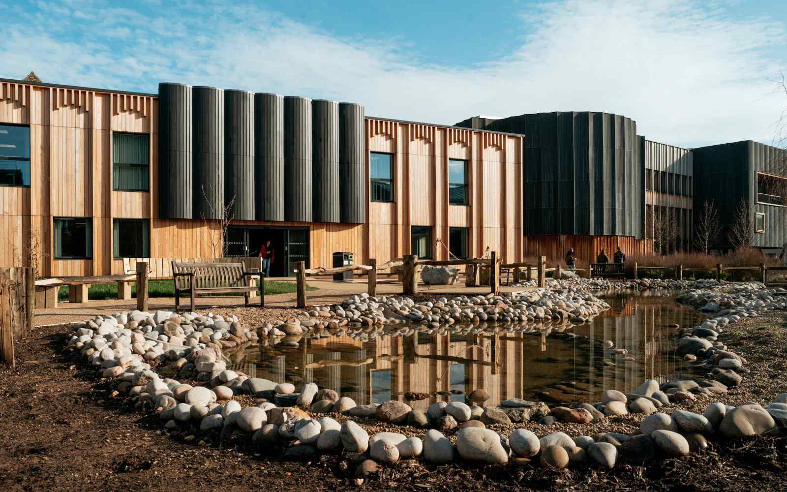 A large timber clad building with a wildlife pond in the foreground