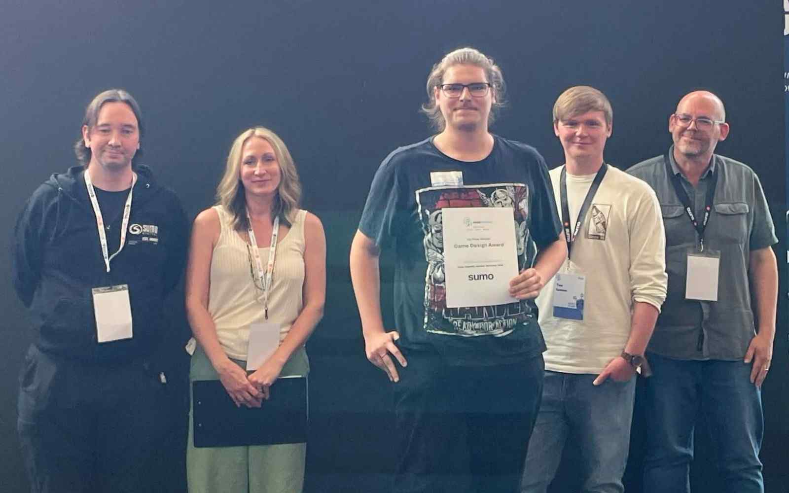 5 people standing in a line looking forwards with the person in the middle holding a certificate