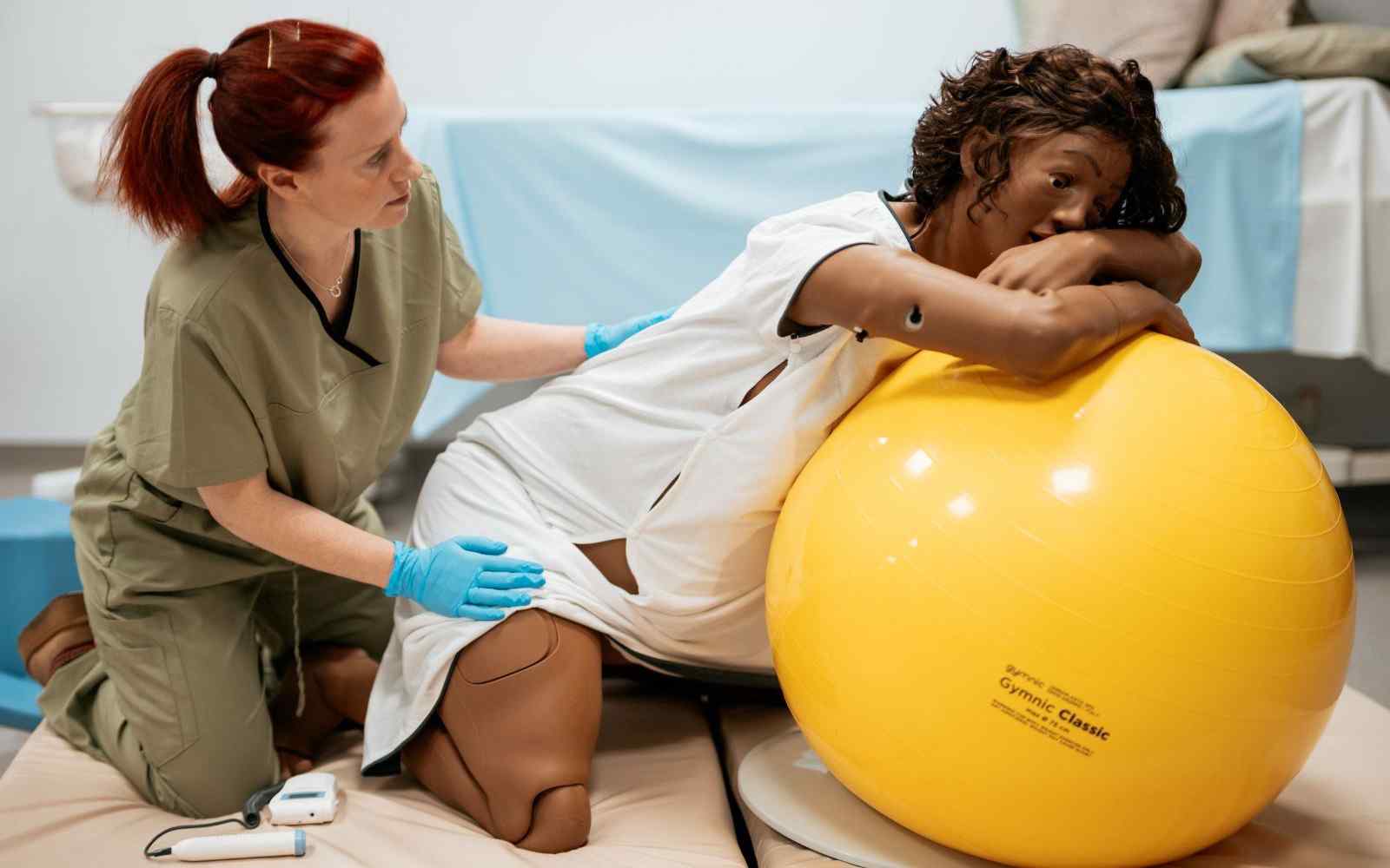 A midwife assisting a manikin birthing mother leaning over a yellow birthing ball
