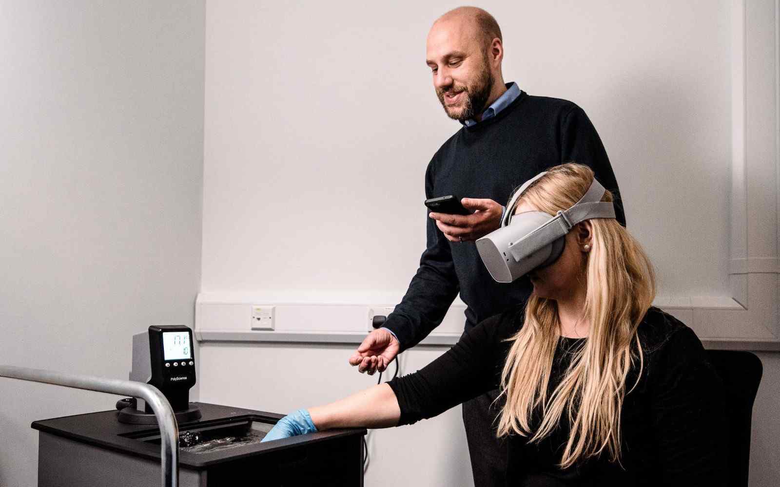 Two people working with VR technology, one seated in a headset with their hand in a waterbath