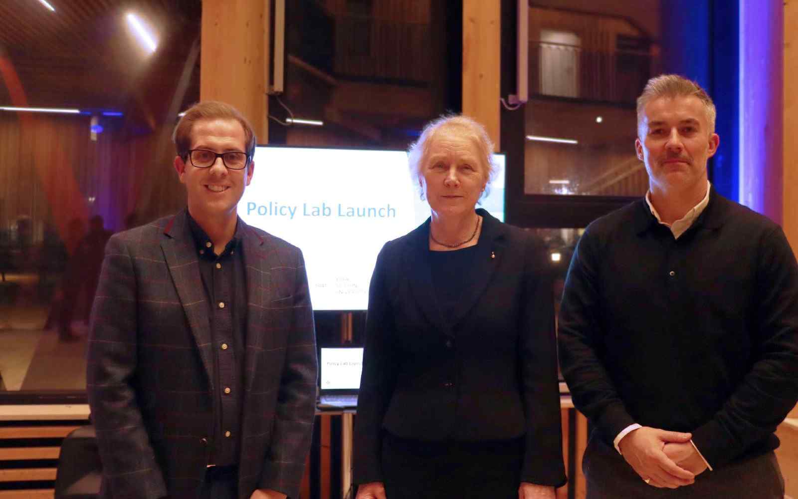 3 people at an event standing in front of an illuminated sign reading Policy Lab Launch