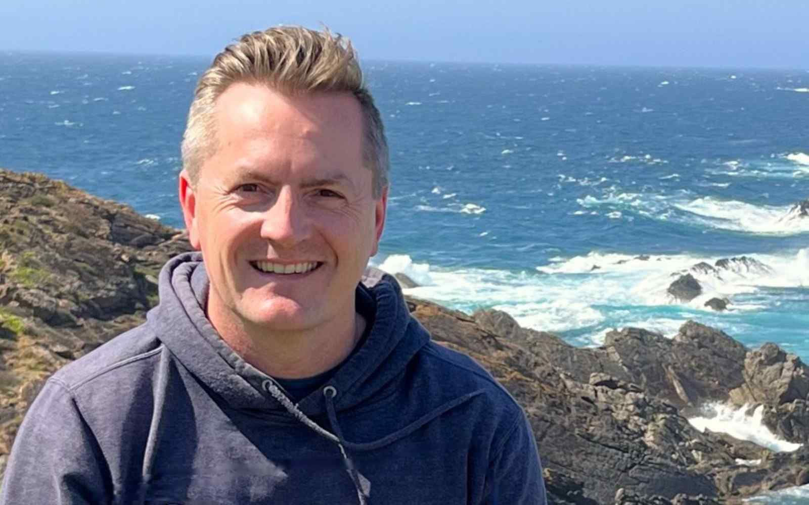 A man pictured in a sunny coast setting with rocks and sea behind him
