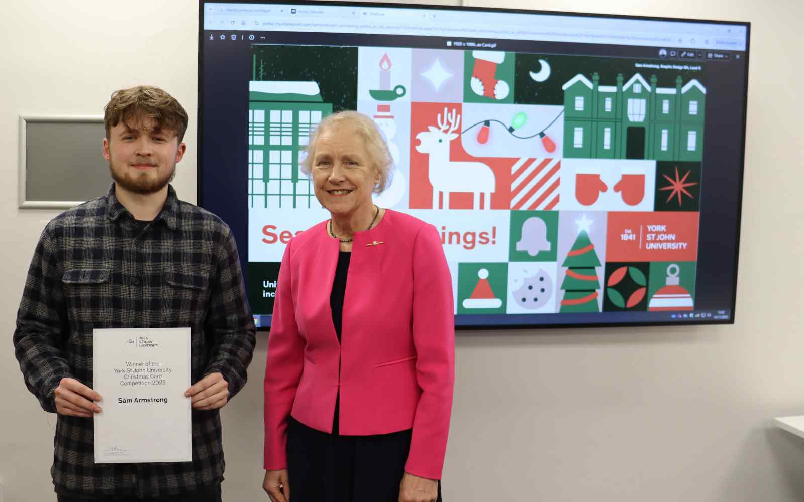 Two people stood in front of a large screen with christmas images on. One person is holding a certificate.