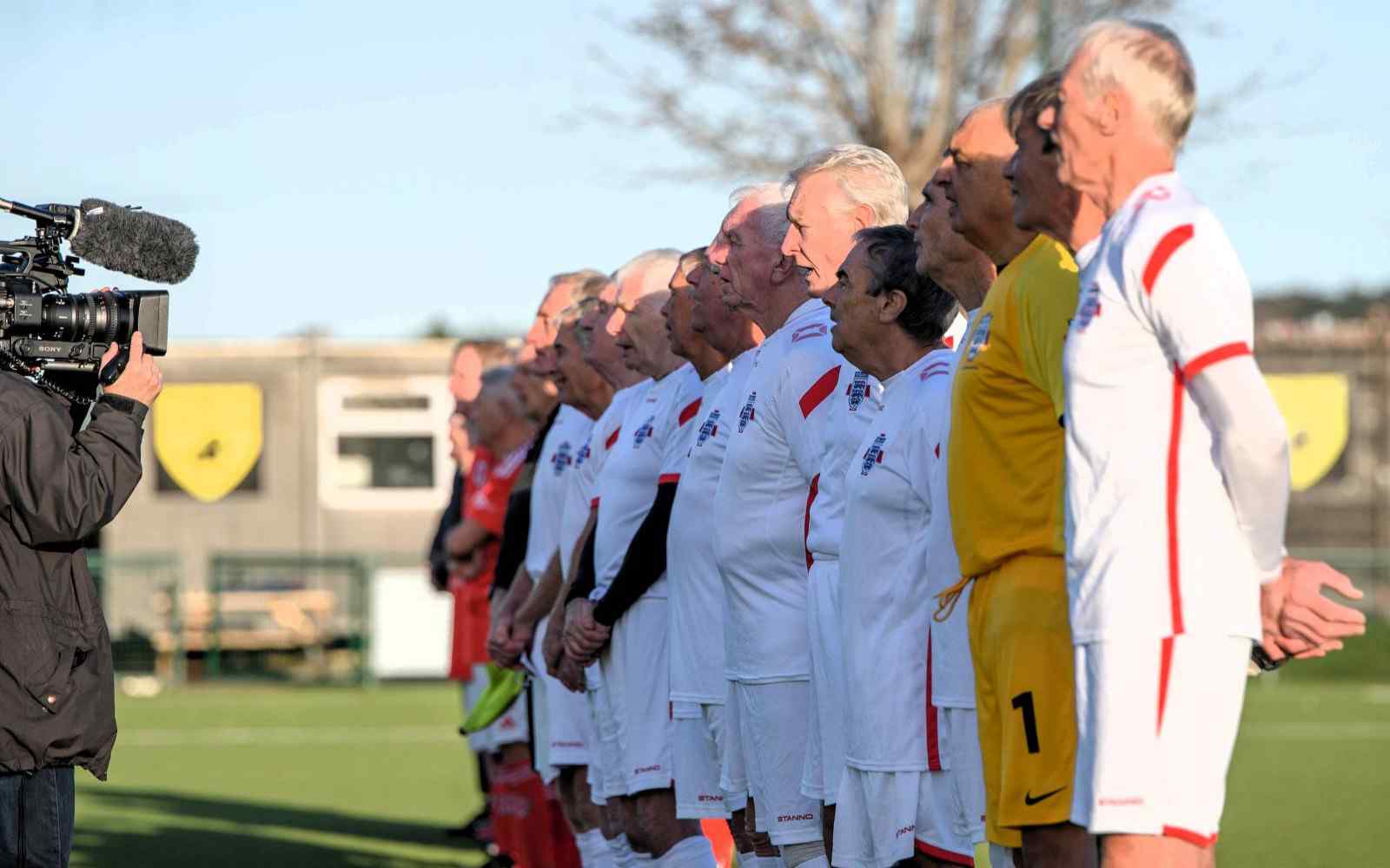 A row of older footballers in England kit being filmed with a TV camera