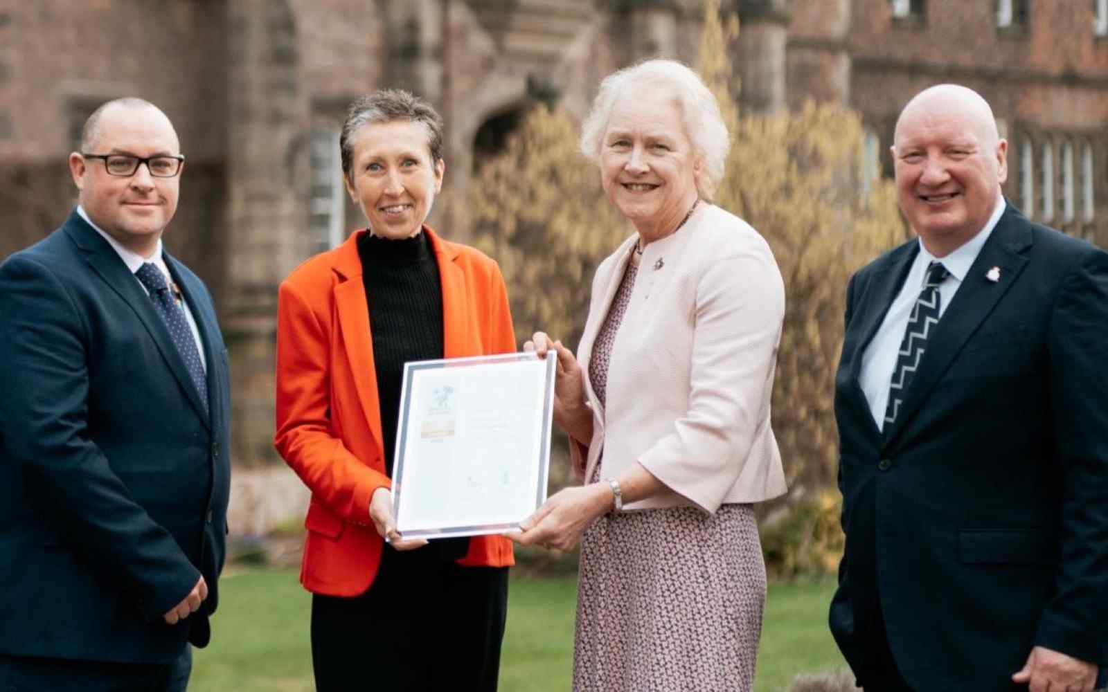 4 people at the hand over a certificate outside the York St John University main entrance
