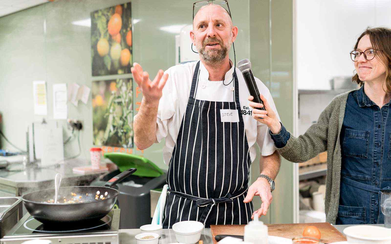 Chef giving cooking demonstration with person holding microphone for them.