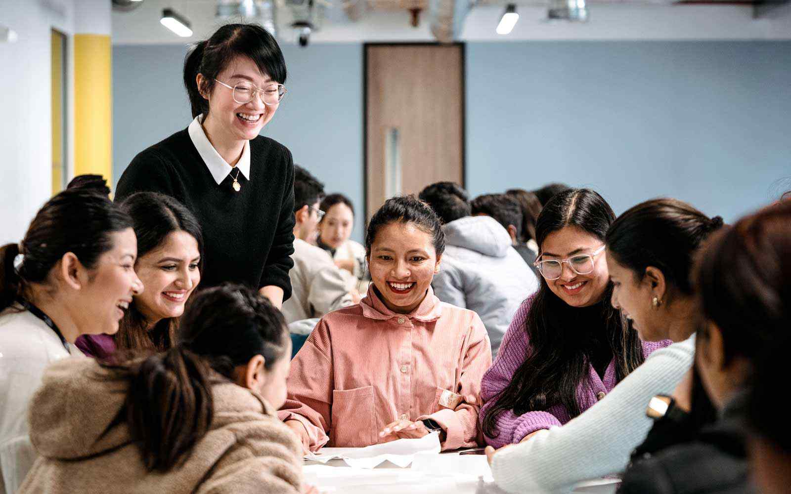 Lecturer talking to group of students gathered around table, working on large sheets of paper.