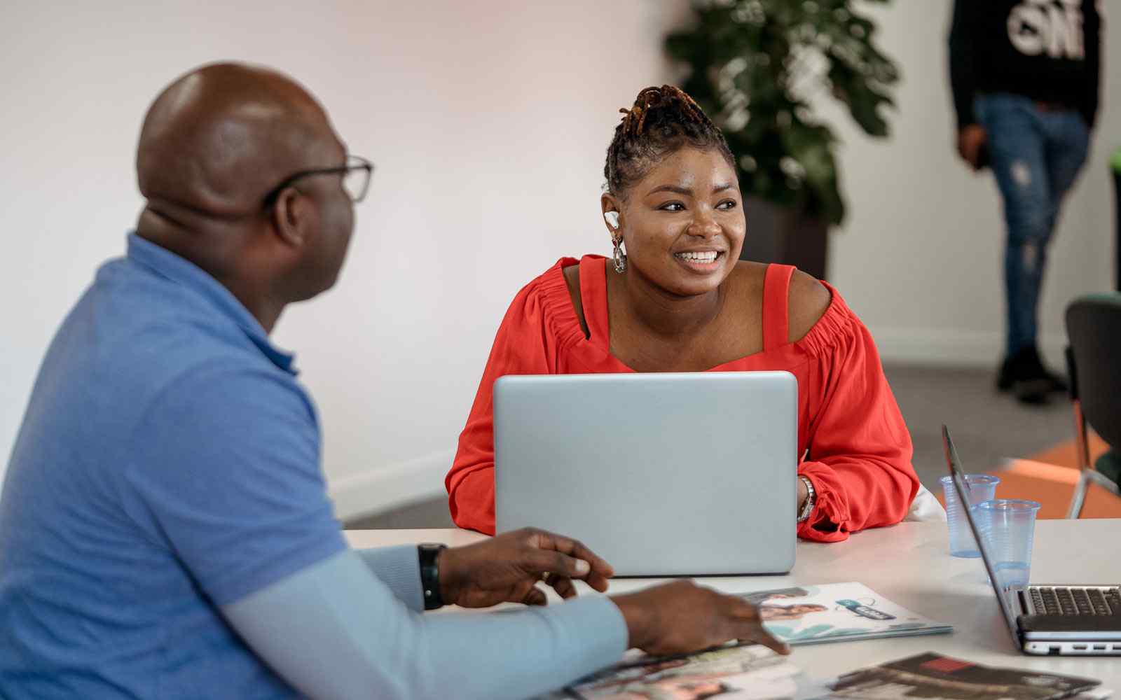 Two people sat at work desk in discussion. One person is using a laptop.