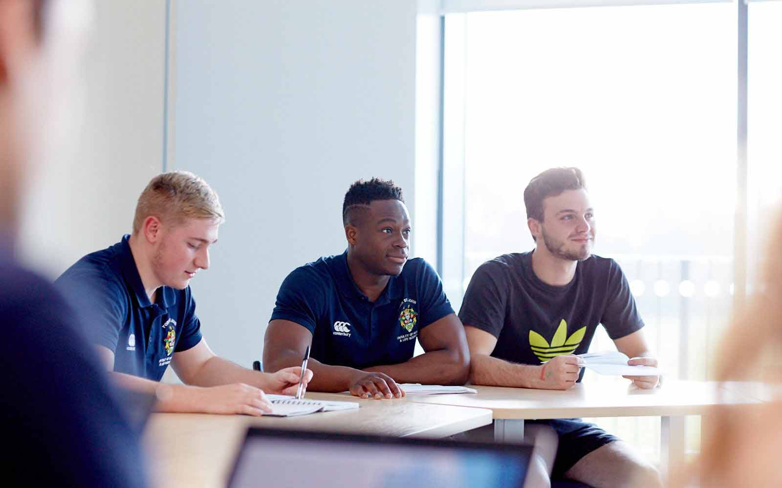 three students sitting at a desk. Stationery is also on the desk