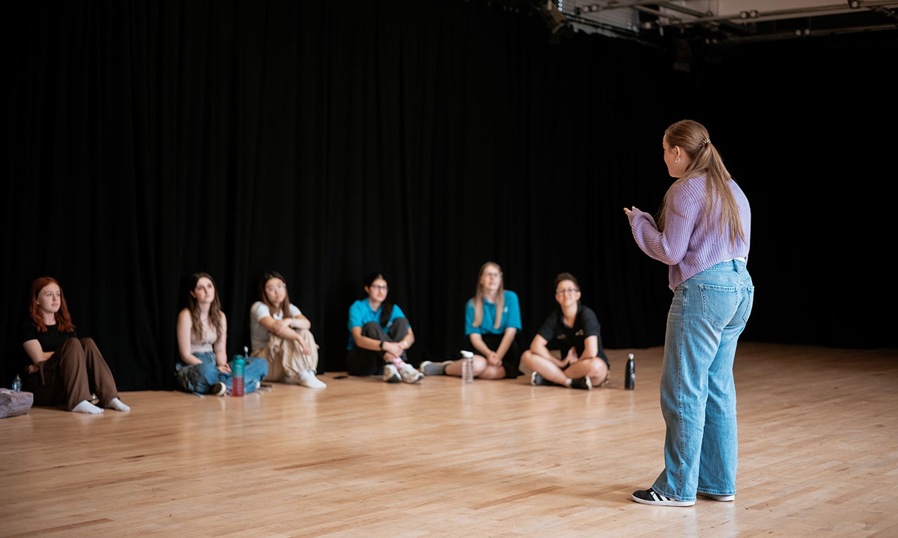 Tutor stood in front of students sitting on floor in drama studio 