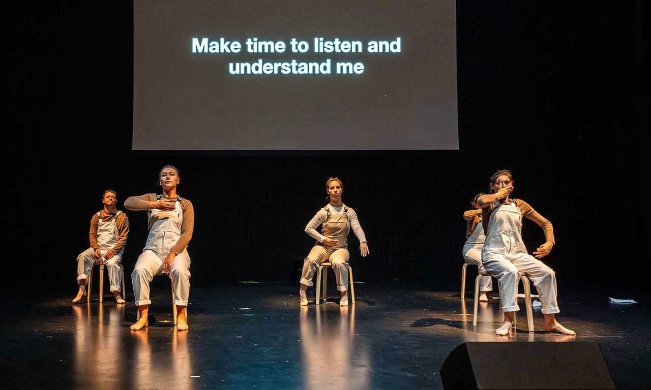 Performers sit on chairs while the words 'Make time to listen and understand me' are projected behind them. 