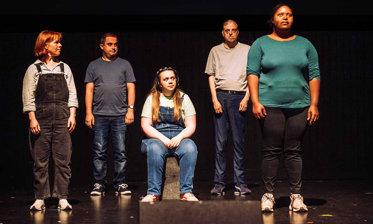 Performers stand in a line, facing out to audience from stage. 