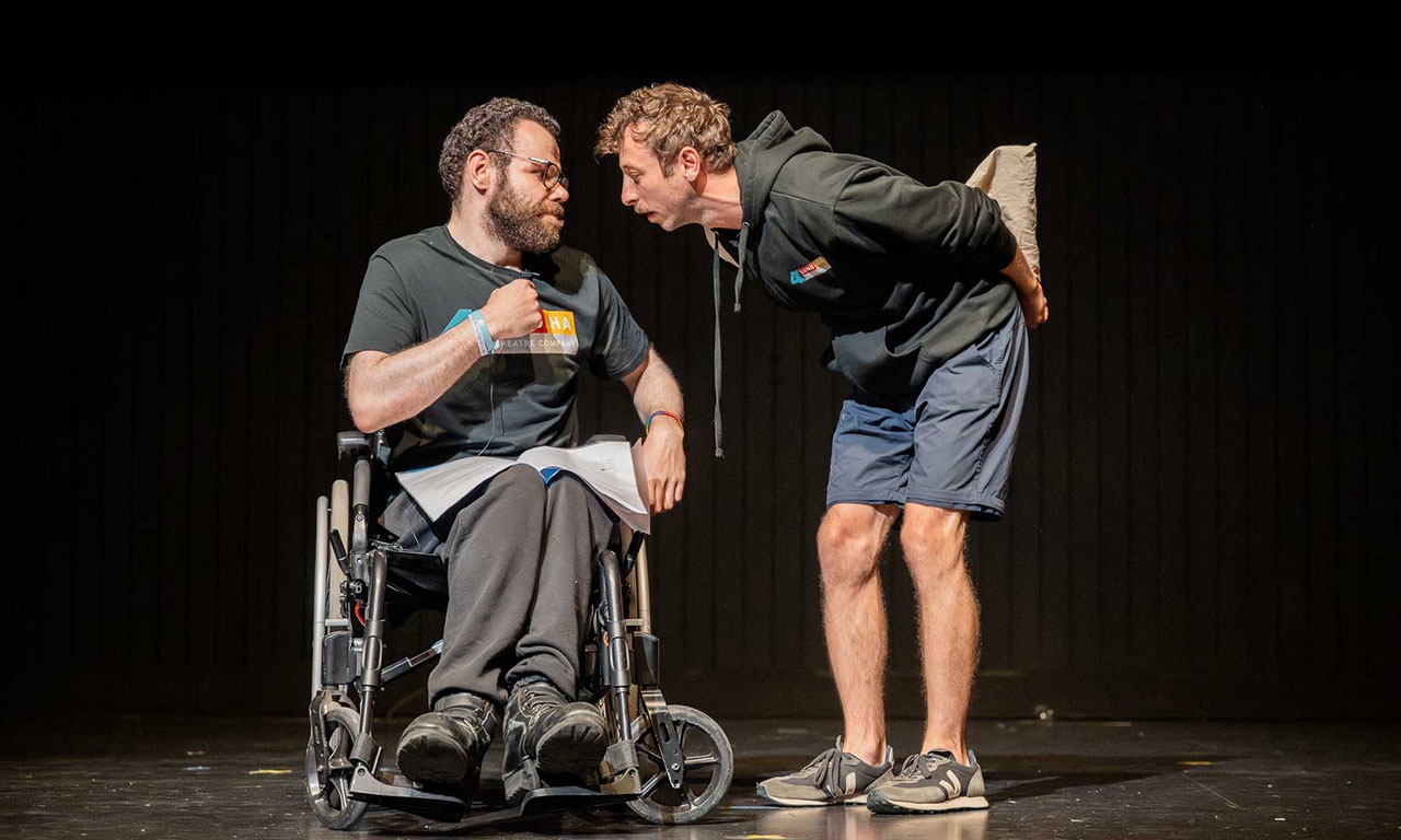Performers on stage. One performer is in a wheelchair while the other bends down to talk to them. 