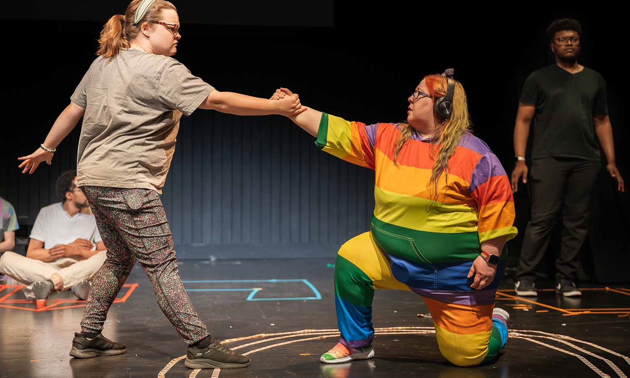 Two performers on stage, one kneeling down and holding the hand of the other who is standing. 
