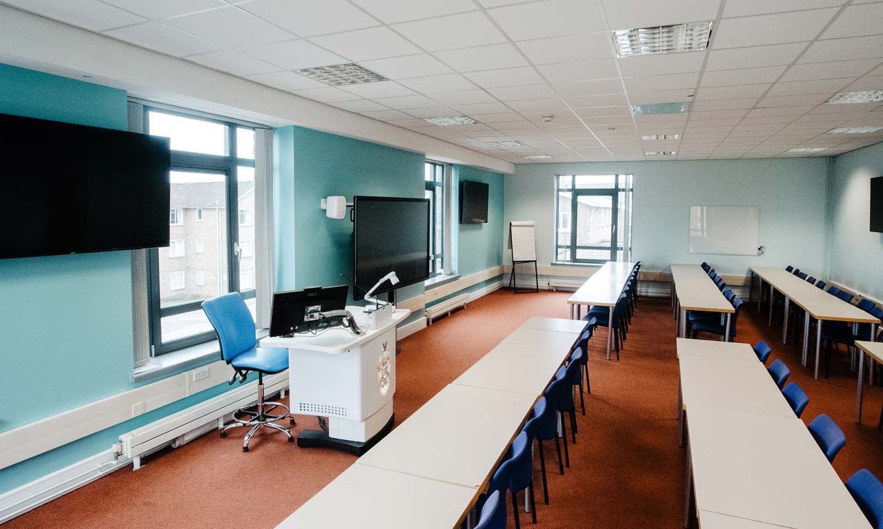 Seminar room with long rows of tables and chairs facing front. Screen and desk at front of room. 