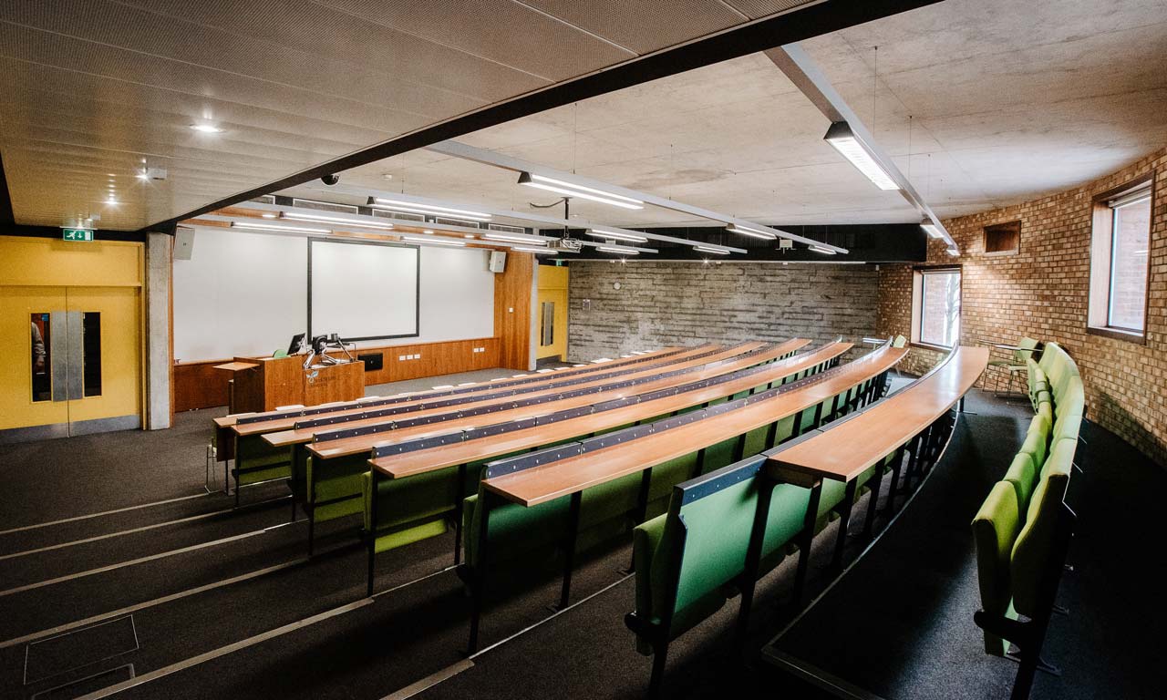 Lecture room looking from back to front. Tiered seating with desk area at front of room. 