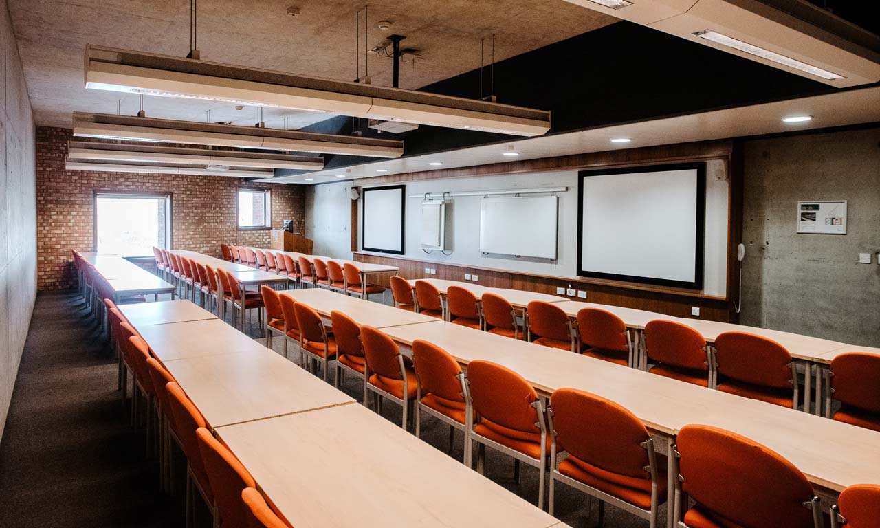 Seminar room with long rows of tables and chairs facing front. Screen and desk at front of room. 