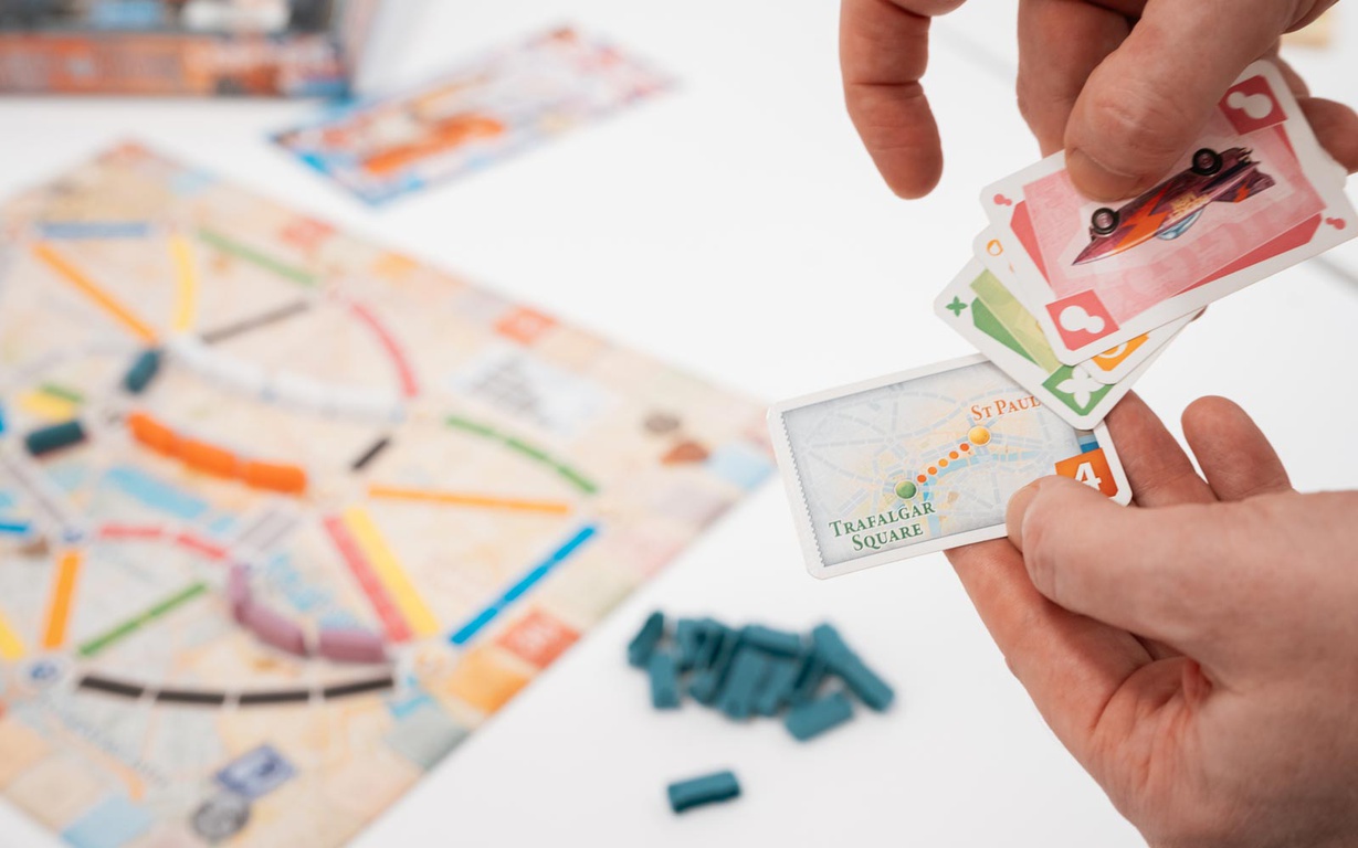 A pair of hands holding cards for the game Ticket to Ride, with the game board in the background. 