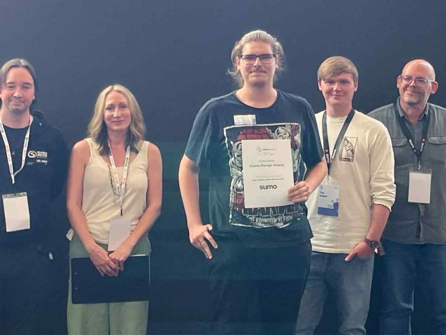 5 people standing in a line looking forwards with the person in the middle holding a certificate 