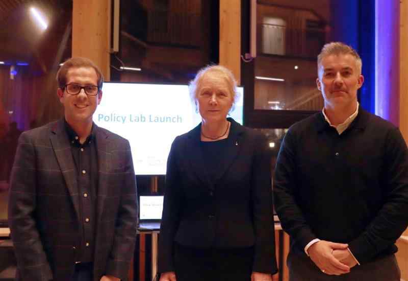 3 people at an event standing in front of an illuminated sign reading Policy Lab Launch 