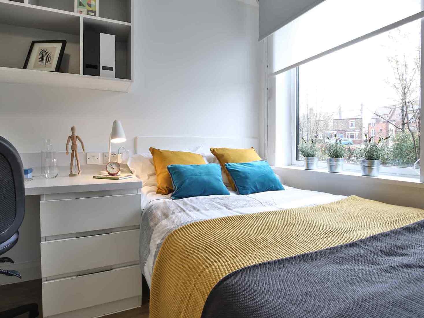 A student room featuring a double bed next to a window, a desk, and some shelves. 
