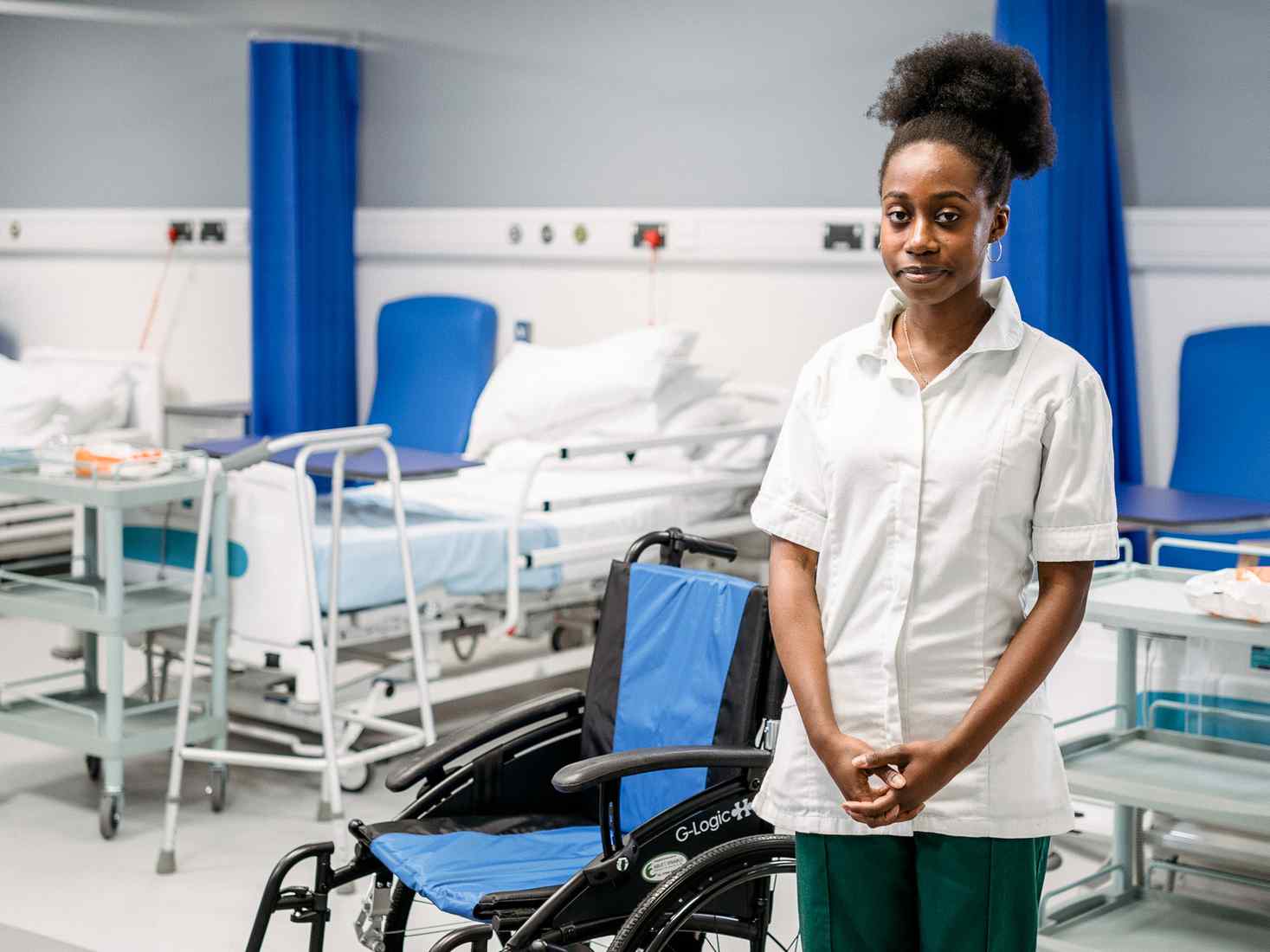 A student on a hospital ward, stood in front of a bed and a wheelchair with their arms folded 