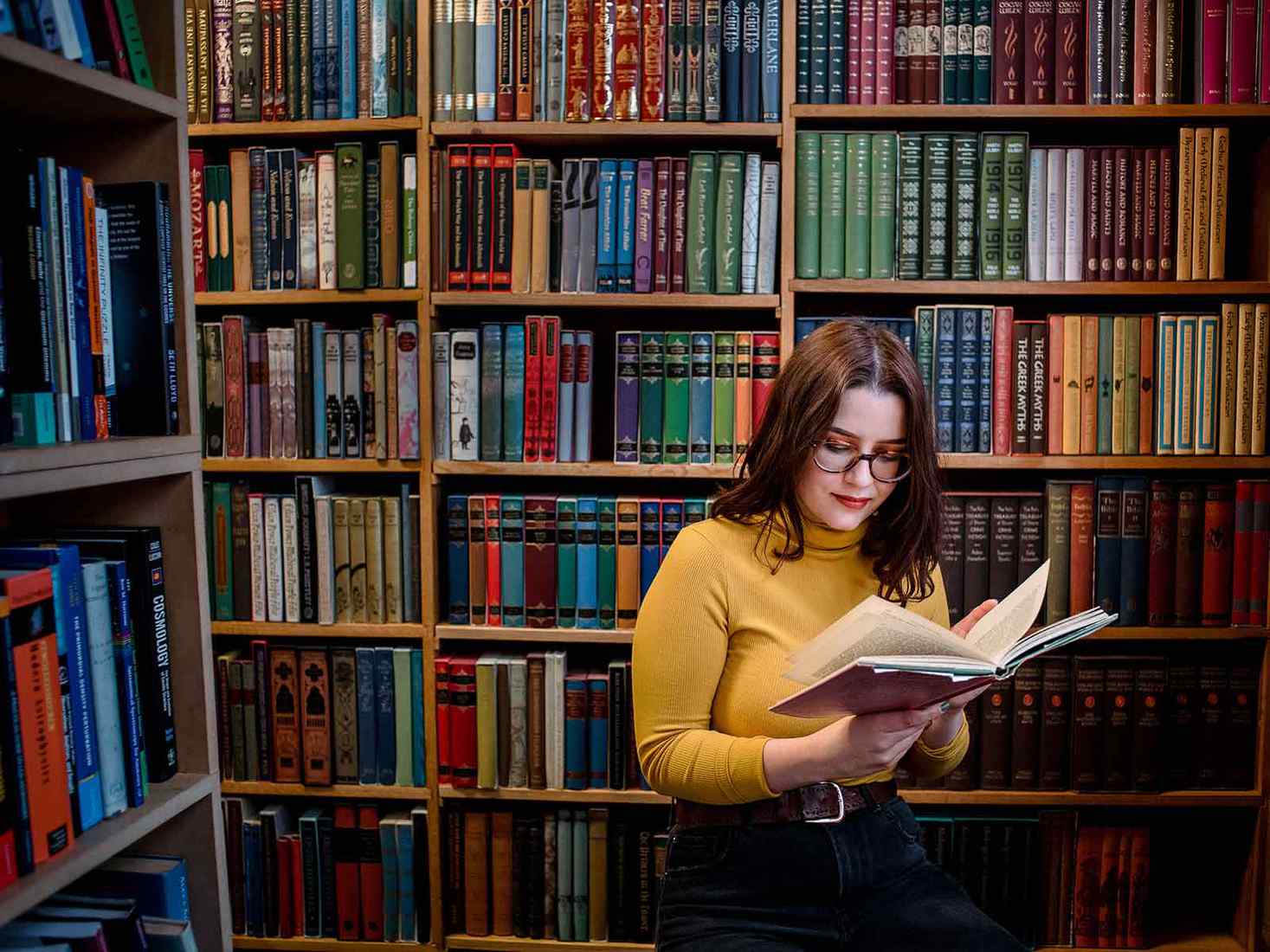 Student reading behind her are lots of books. She is wearing a blue jumper and jeans 