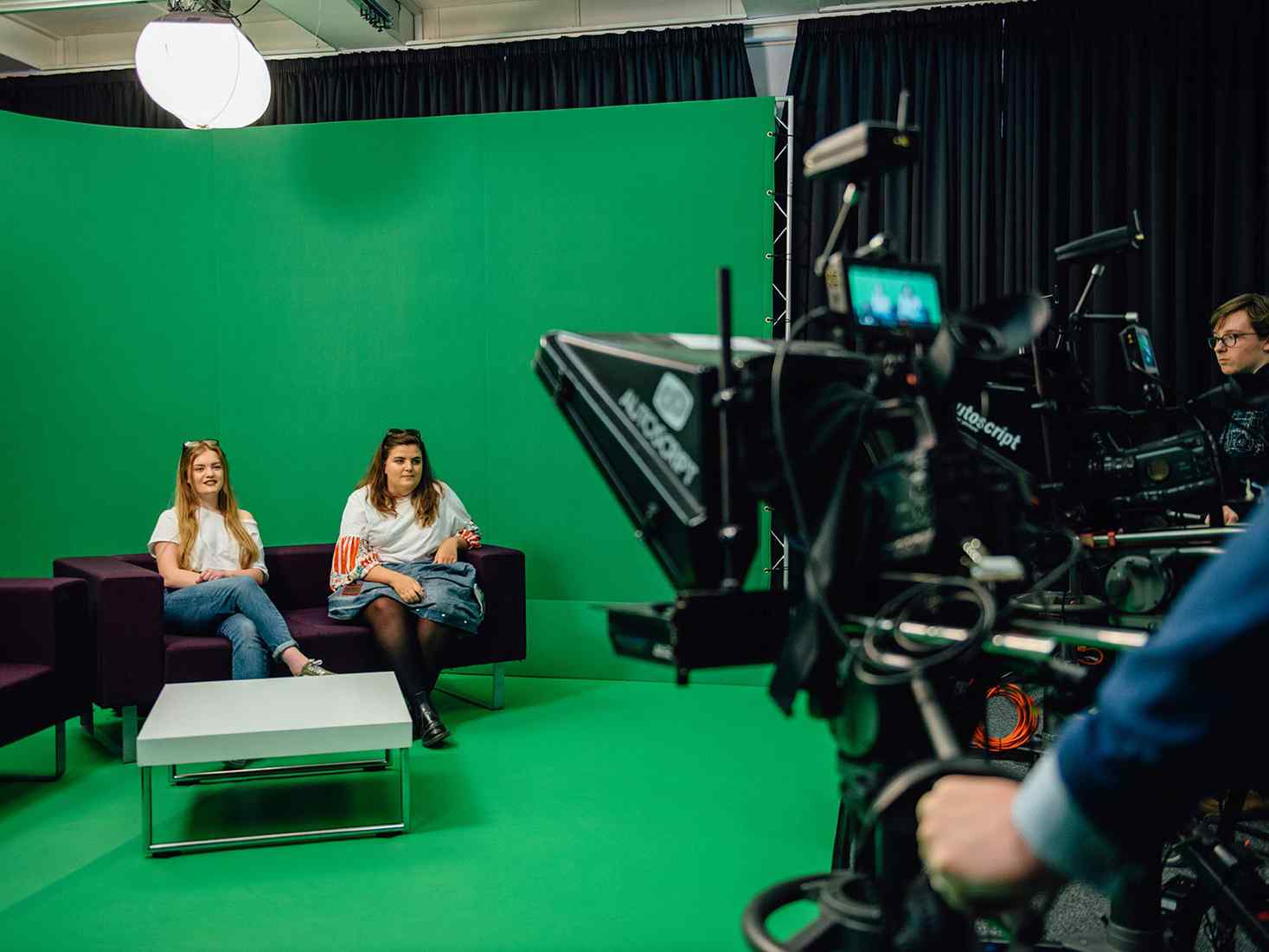 Two students sat in front of a green screen 