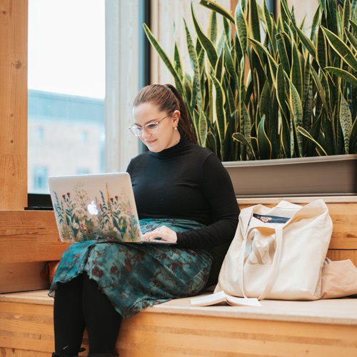 A student sitting on a bench in the Creative Centre with their laptop. 